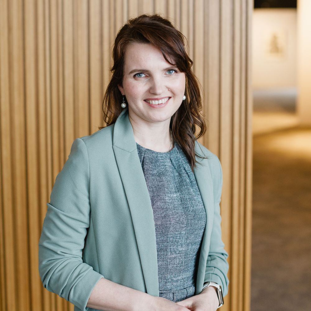 Woman in a light blue blazer standing in front of a wooden paneled wall.