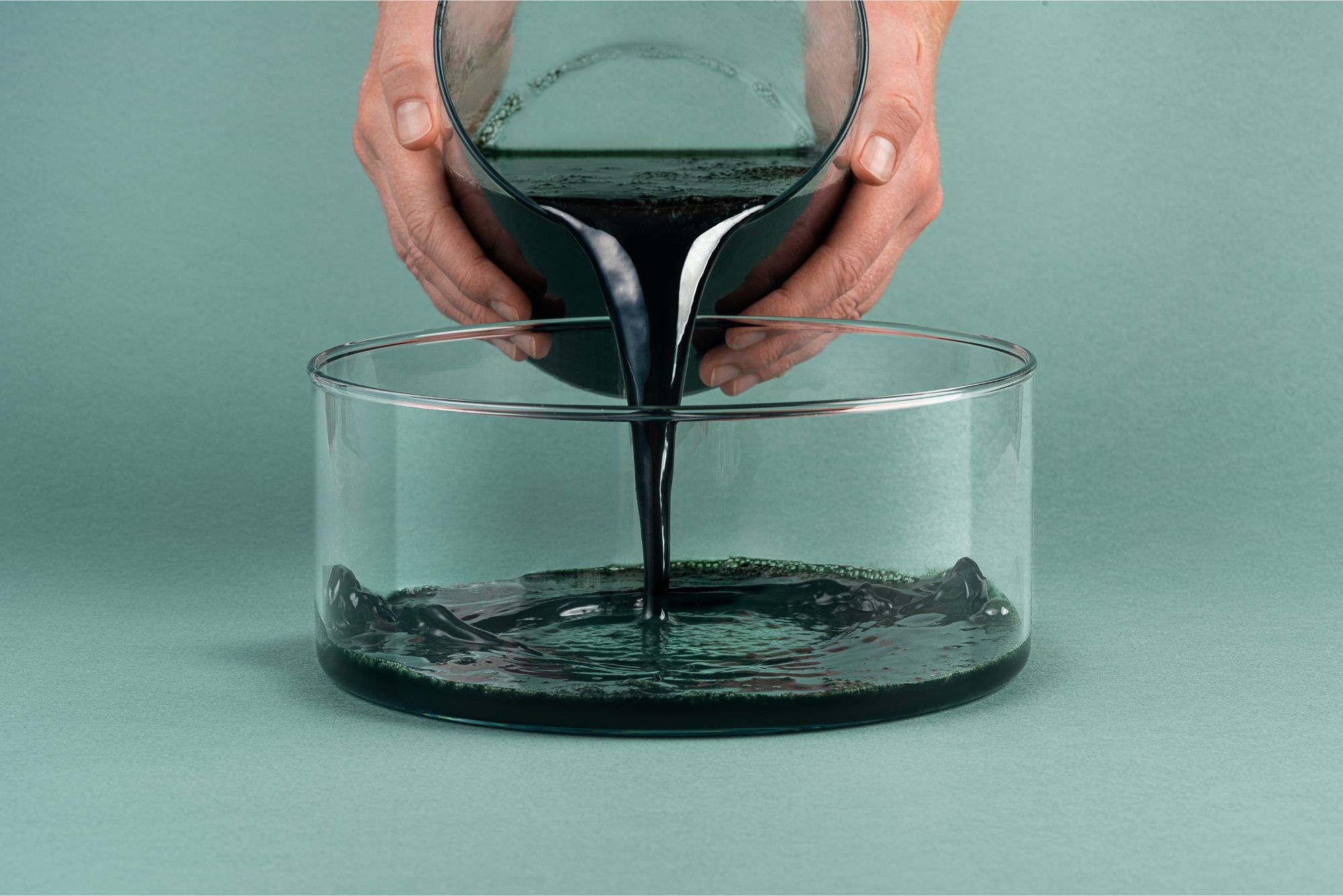 Person pouring raw and living spirulina from a glass container into a larger glass bowl on a green background